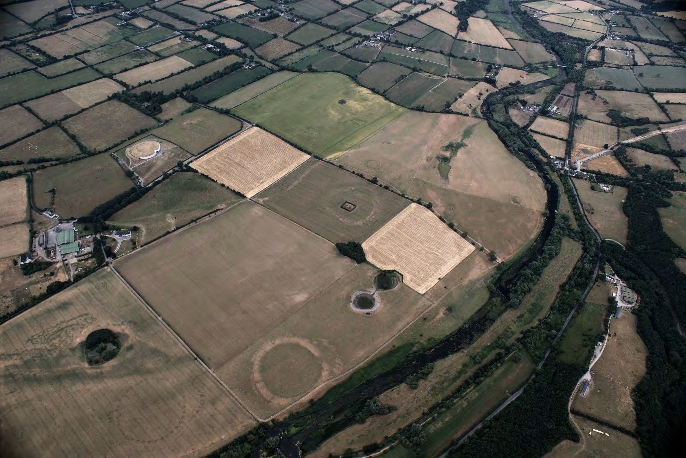 Oblique aerial view of the Newgrange floodplain from the south-west, with the river, pale pasture and circular marks in the fields.