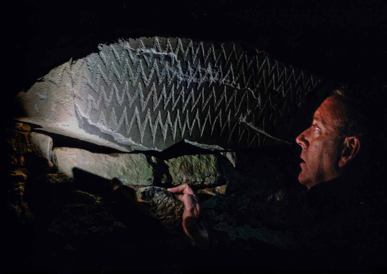 Anthony Murphy in profile inside a dark passage tomb, looking at a stone carved with zigzag lines, lit by a torch.