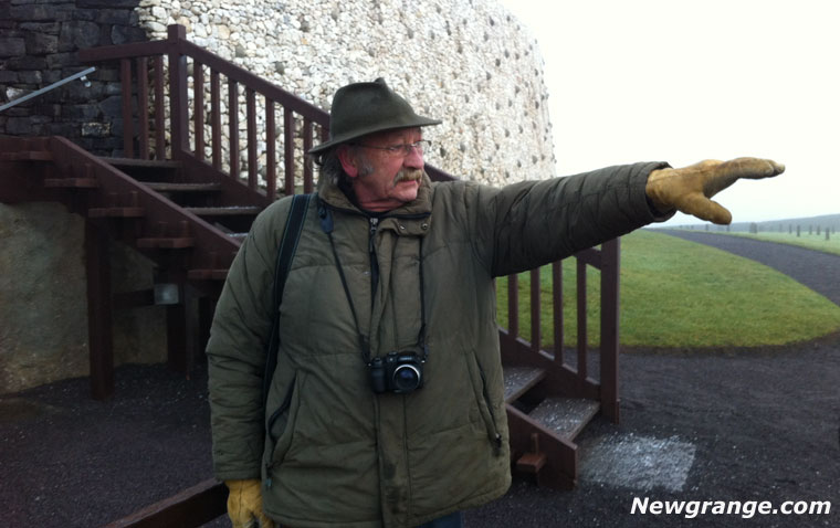 Jack Roberts in a hat and winter coat pointing across the Newgrange mound, a camera around his neck, white quartz wall and wooden stairs behind.