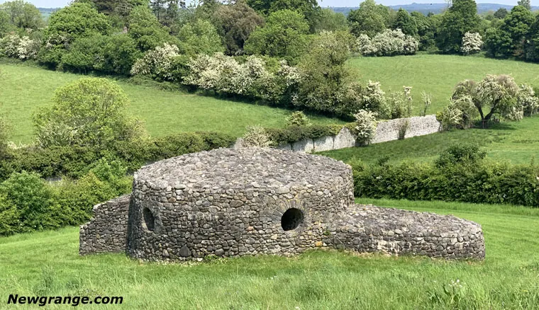 Circular dry-stone Newgrange folly with two round openings, set in a grassy field with hedges and trees behind.