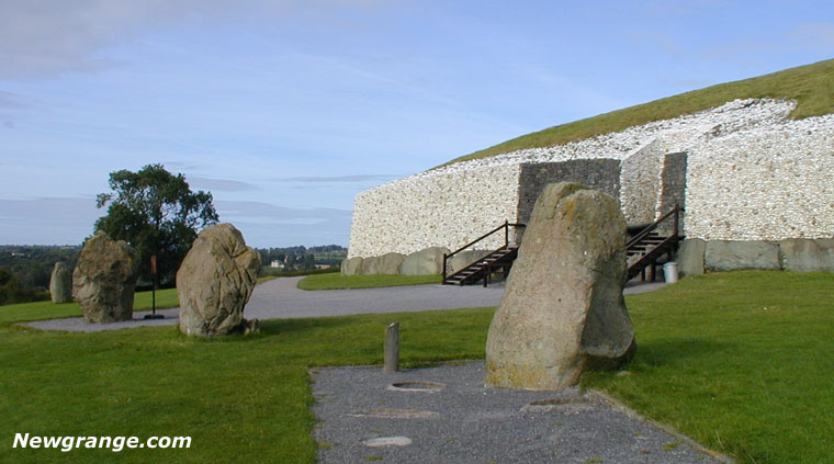 Great Circle standing stones on grass before the white quartz facade at Newgrange, with wooden stairs to the entrance.
