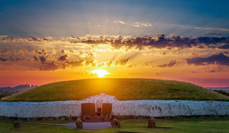 Newgrange at sunrise with sun behind the mound, crepuscular rays through clouds, kerbstones, roof box and entrance orthostats visible